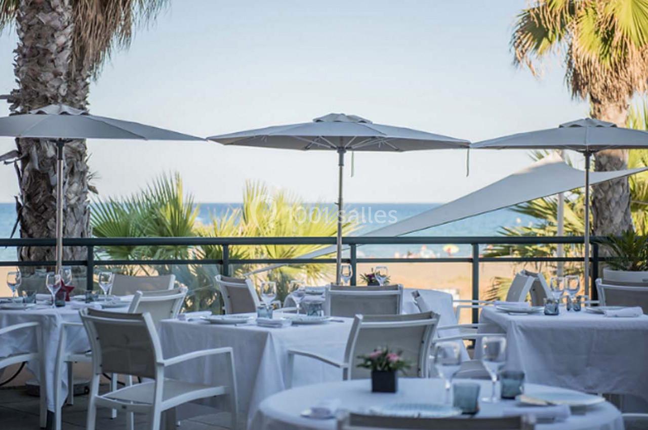 Terrasse d'un restaurant avec tables dressées, vue sur la plage, palmiers et mer en arrière-plan.