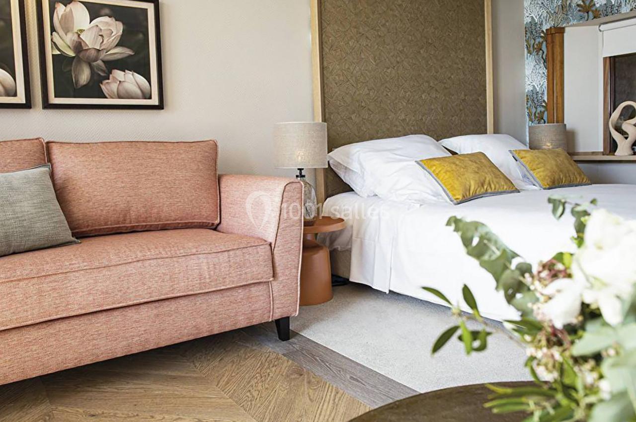 Chambre lumineuse avec un canapé rose, un lit double aux coussins jaunes et une table décorée de fleurs.