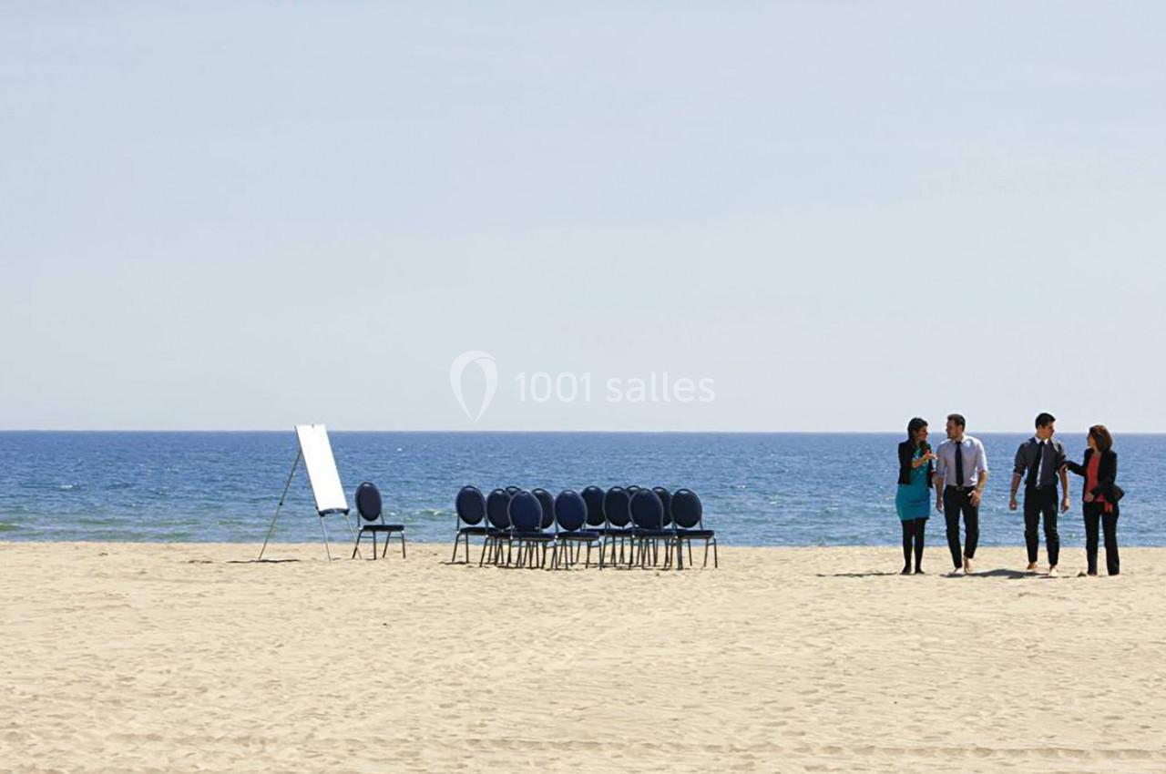 Un groupe de personnes marche sur une plage près de chaises alignées et d'un tableau blanc face à la mer.