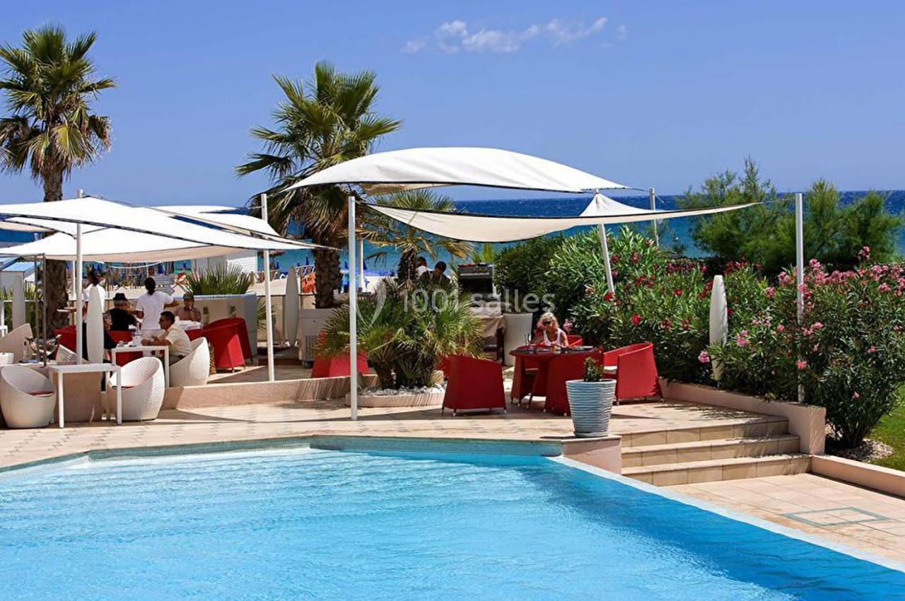 Terrasse en bord de piscine avec parasols blancs, mobilier rouge et vue sur la mer entourée de végétation.
