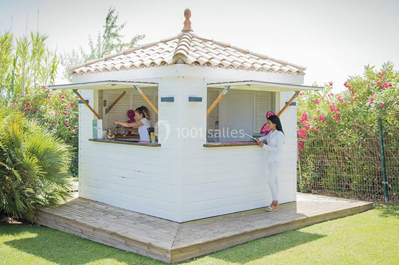 Kiosque en bois blanc avec toit en tuiles, entouré de végétation, utilisé comme espace de service extérieur.