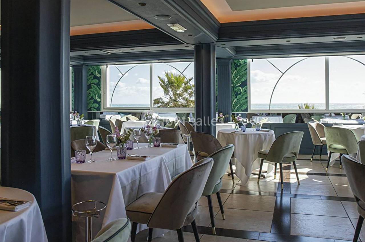 Salle de restaurant lumineuse avec des tables dressées, grandes baies vitrées et vue sur la mer et des palmiers.