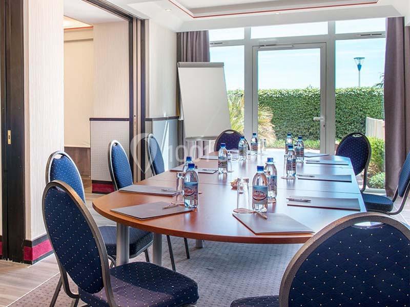 Salle de réunion lumineuse avec table ovale, chaises bleues, bouteilles d'eau, bloc-notes et tableau de présentation.