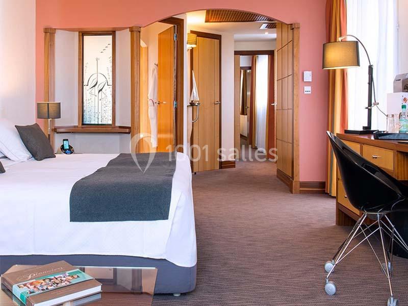 Chambre d'hôtel lumineuse avec lit double, bureau, chaise, et couloir menant à une salle de bain.