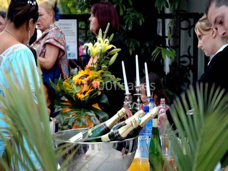 Des bouteilles de champagne dans un seau à glace sur une table, entourées de fleurs et de convives en arrière-plan.