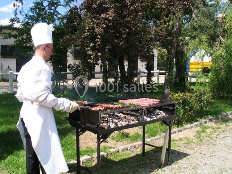 Un chef en tenue blanche cuisine des brochettes et viandes sur un barbecue en plein air, entouré de verdure.