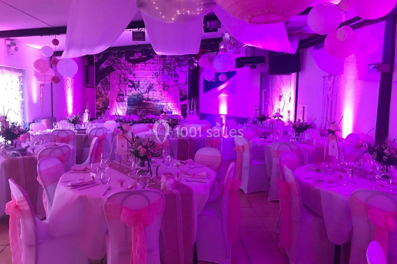 Salle de réception décorée avec des nappes blanches, des rubans roses et un éclairage d'ambiance violet.