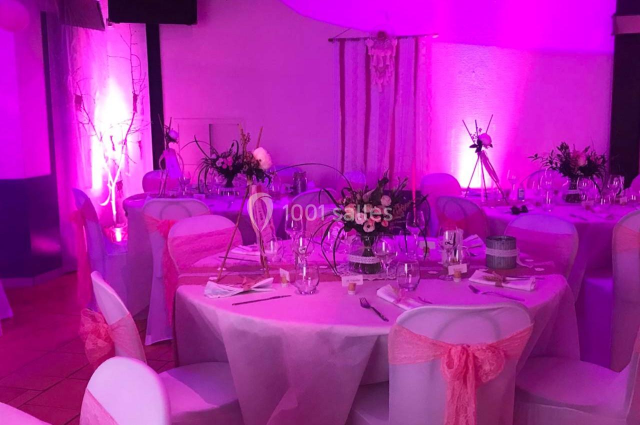 Salle décorée pour un événement, avec tables rondes ornées de nappes roses, chaises habillées et éclairage tamisé rose.