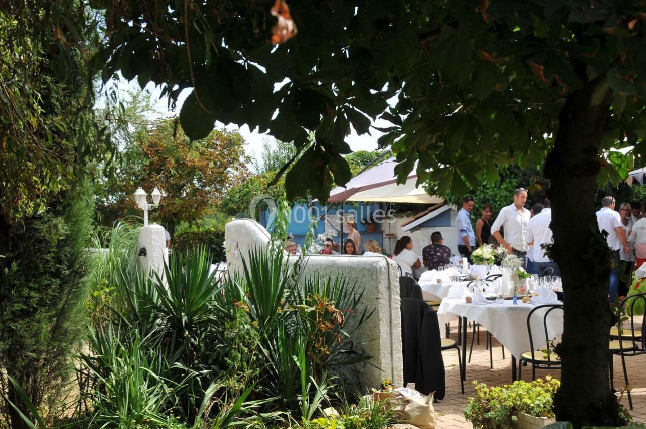 Jardin ombragé avec des tables dressées pour un repas en plein air, entouré de végétation et de convives.