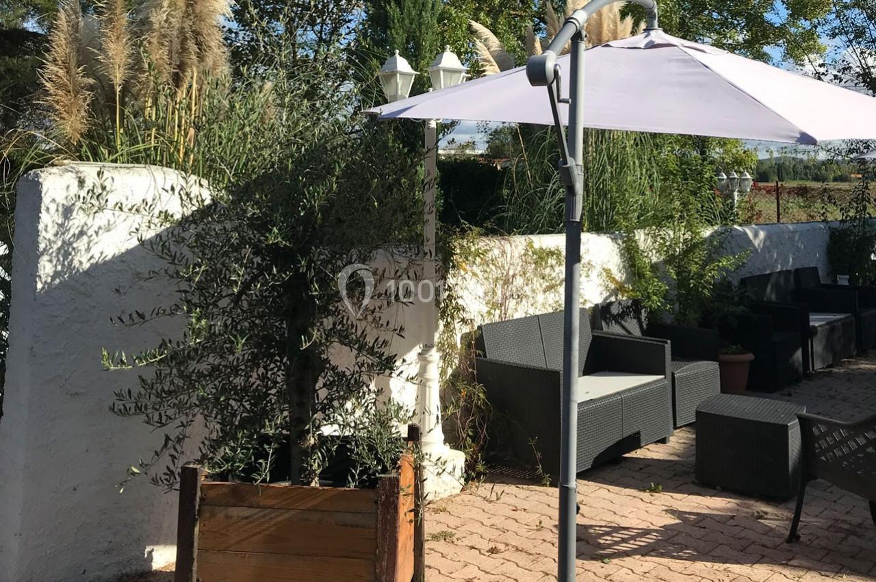 Terrasse ensoleillée avec parasol blanc, mobilier de jardin en résine tressée et végétation en arrière-plan.