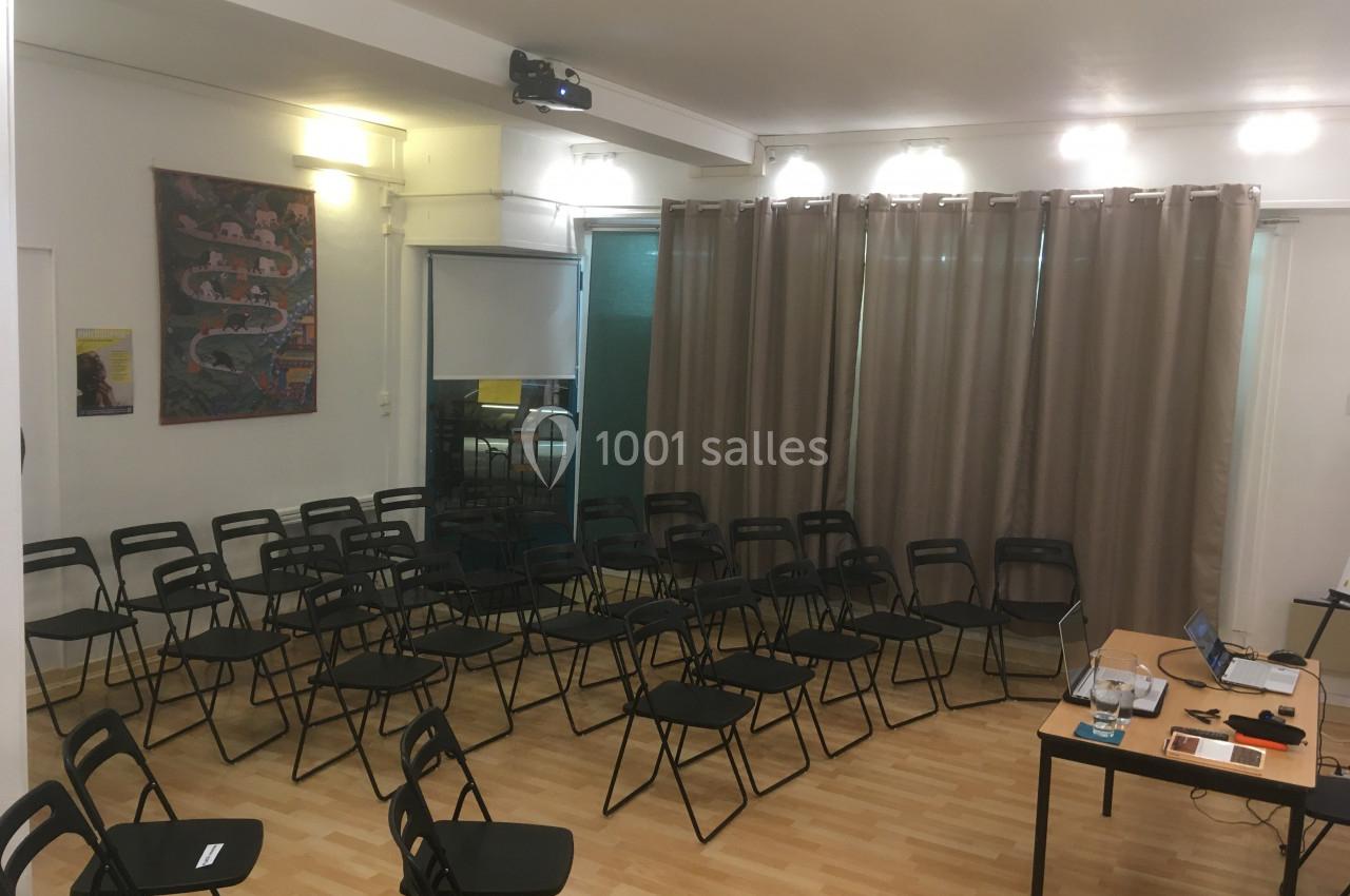 Salle lumineuse avec des chaises noires disposées en rangées, un bureau avec matériel et un projecteur au plafond.