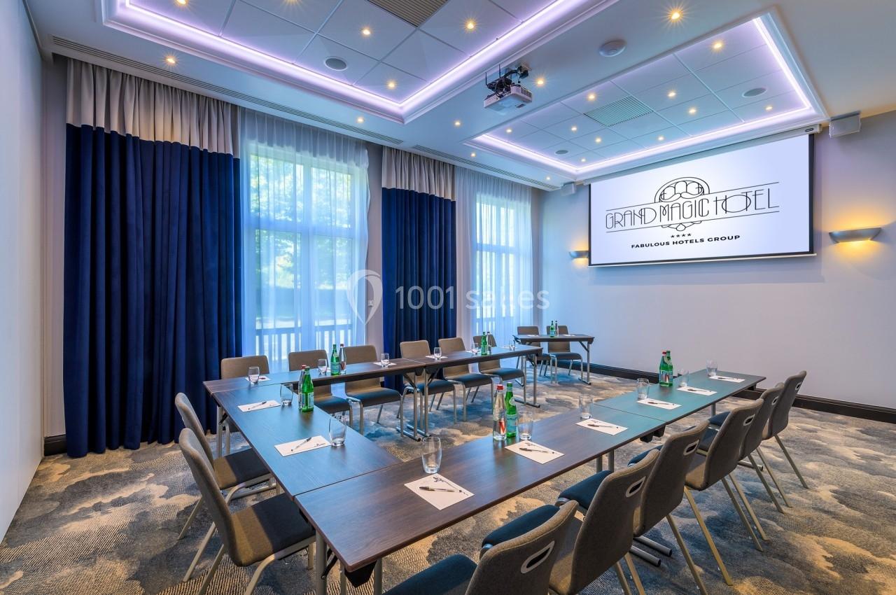 Salle de réunion lumineuse avec tables disposées en U, chaises, bouteilles d'eau et écran de projection allumé.