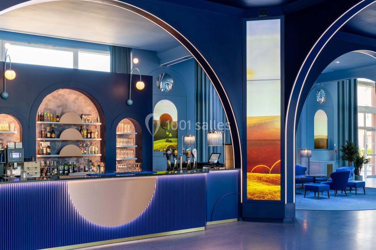Bar moderne avec comptoir éclairé, arches bleues, étagères de bouteilles et décorations lumineuses dans un espace coloré.