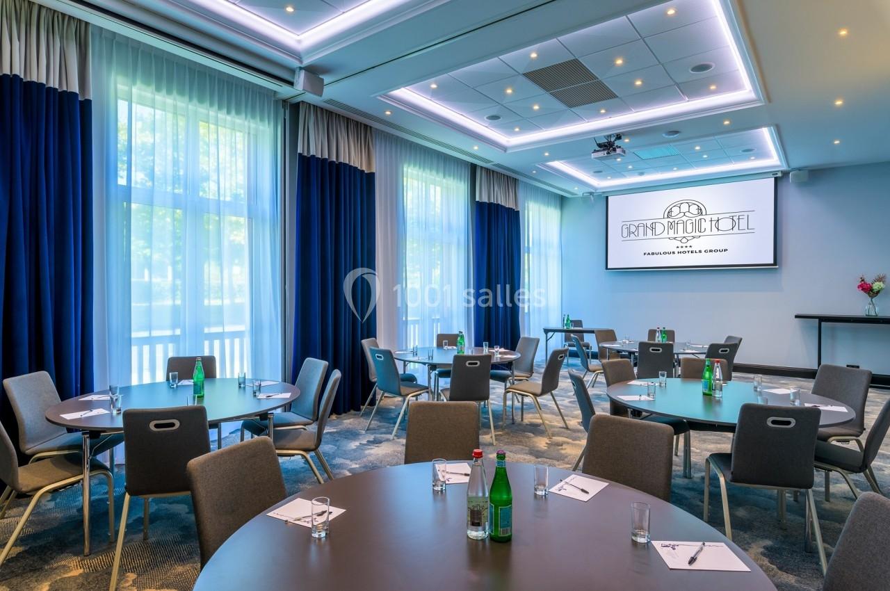 Salle de réunion lumineuse avec tables rondes, chaises, bouteilles d'eau et écran de projection allumé.