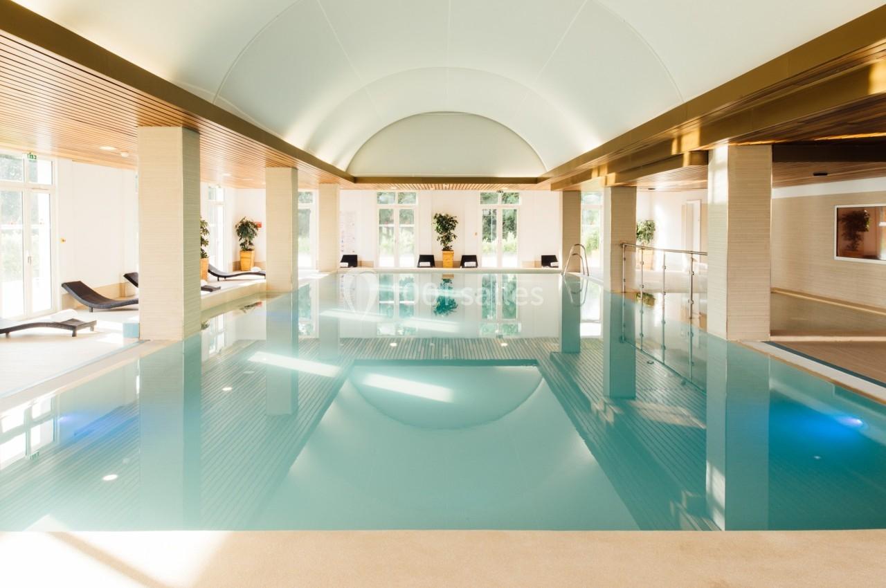 Piscine intérieure lumineuse avec plafond voûté, transats alignés et grandes baies vitrées laissant entrer la lumière…