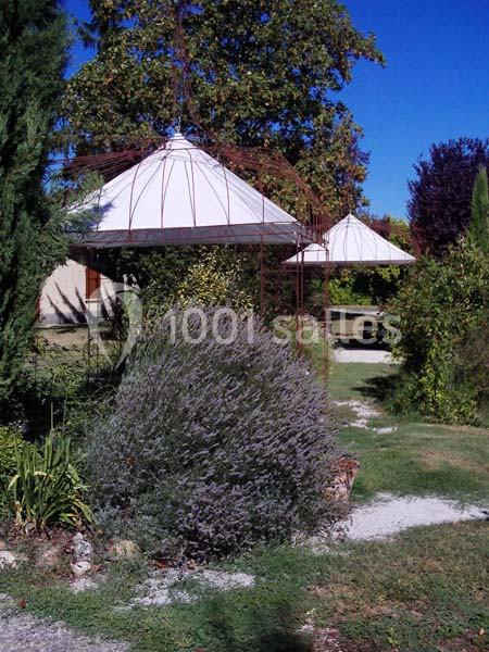 Deux structures métalliques avec toiles blanches dans un jardin arboré, entourées de végétation et d'allées en gravier.