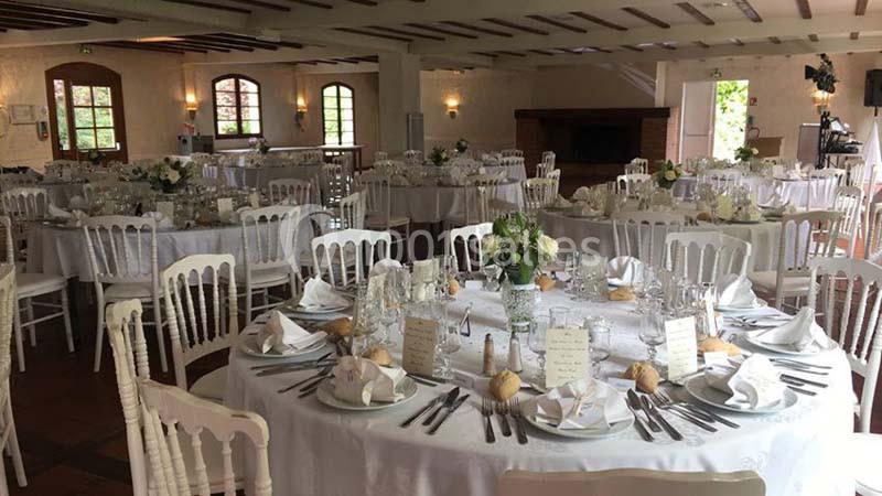 Salle de réception décorée avec des tables rondes dressées, nappes blanches, chaises en bois et arrangements floraux.
