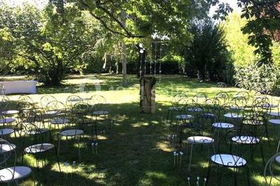 Chaises et tables en fer forgé disposées sous un grand arbre dans un jardin verdoyant, éclairé par la lumière du jour.