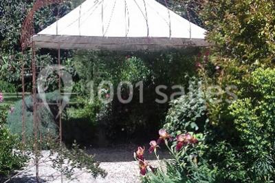 Chaises et tables en fer forgé disposées sous un grand arbre dans un jardin verdoyant, éclairé par la lumière du jour.