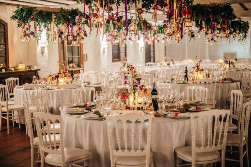 Salle de réception décorée avec des tables rondes, nappes blanches, chandelles et suspensions florales colorées.