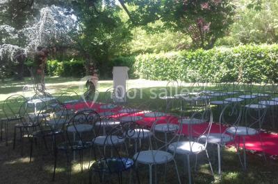 Chaises et tables en fer forgé disposées sous un grand arbre dans un jardin verdoyant, éclairé par la lumière du jour.