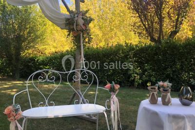 Chaises et tables en fer forgé disposées sous un grand arbre dans un jardin verdoyant, éclairé par la lumière du jour.