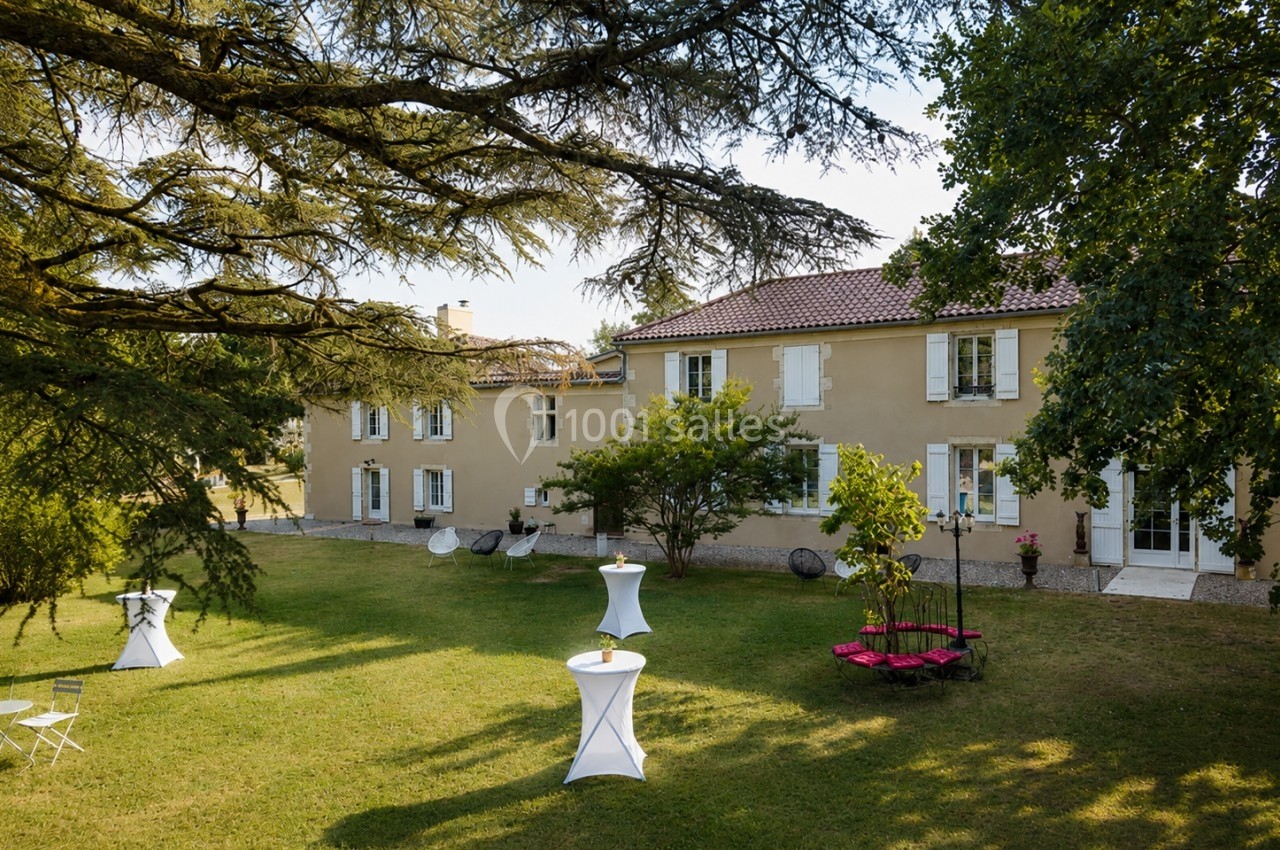 Grande maison en pierre avec volets blancs, entourée d'un jardin arboré et aménagé avec des tables et des chaises.