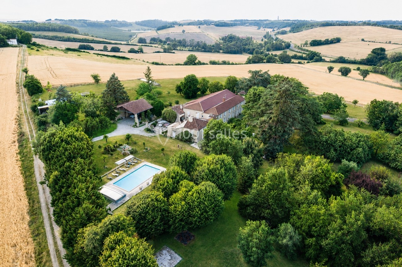 Vue aérienne d'une maison en pierre avec piscine, entourée de jardins verdoyants et de champs vallonnés.
