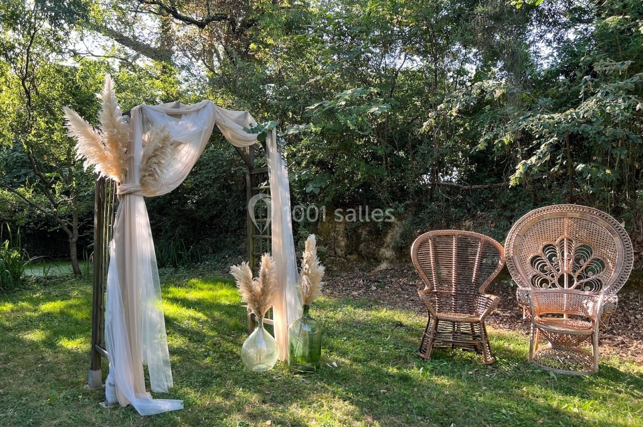 Arche décorée de voilages et de pampas avec deux fauteuils en rotin dans un jardin verdoyant.