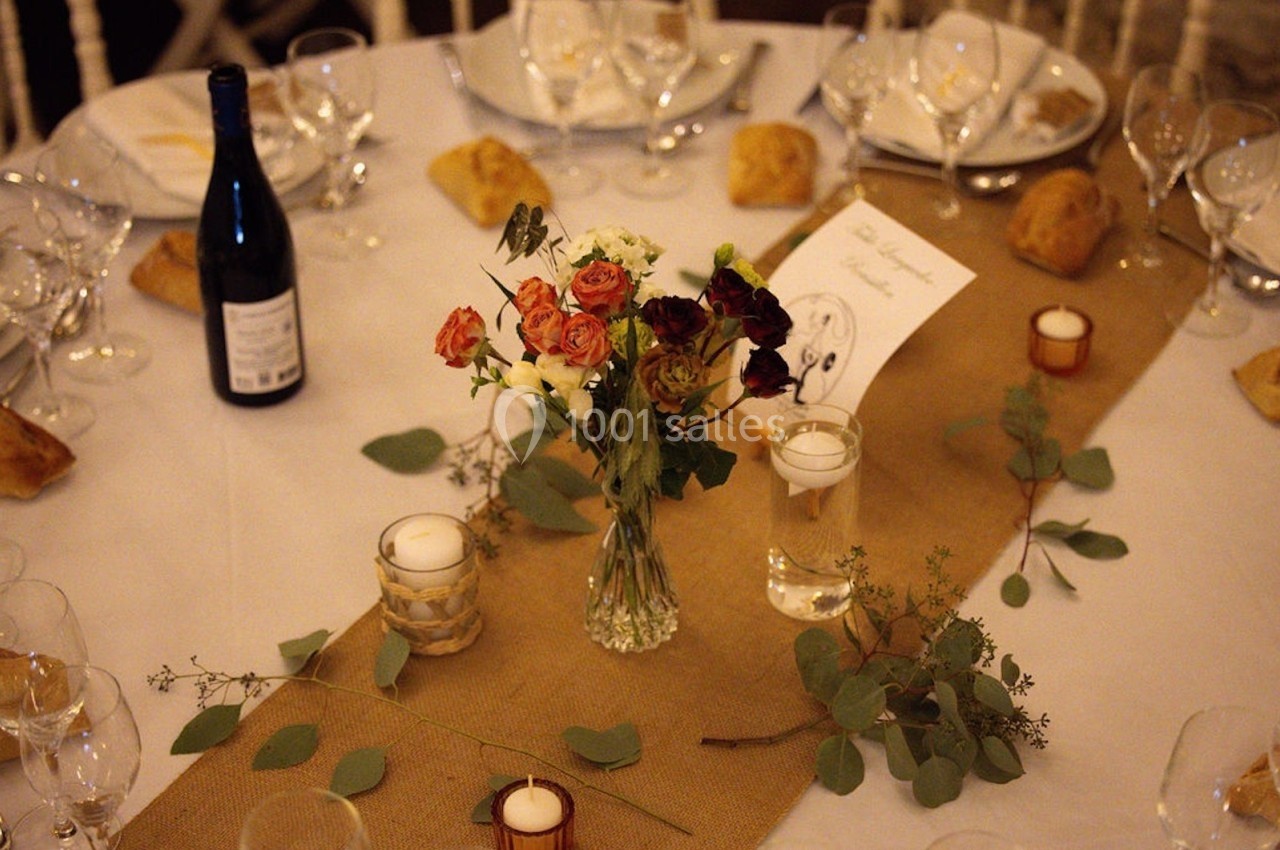 Table décorée avec un chemin de table en jute, un bouquet de fleurs, des bougies, des verres et une bouteille de vin.