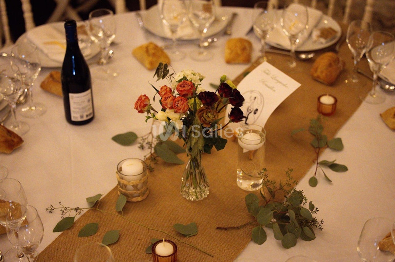 Table décorée avec un chemin de table en jute, un bouquet de fleurs, des bougies, des verres et une bouteille de vin.