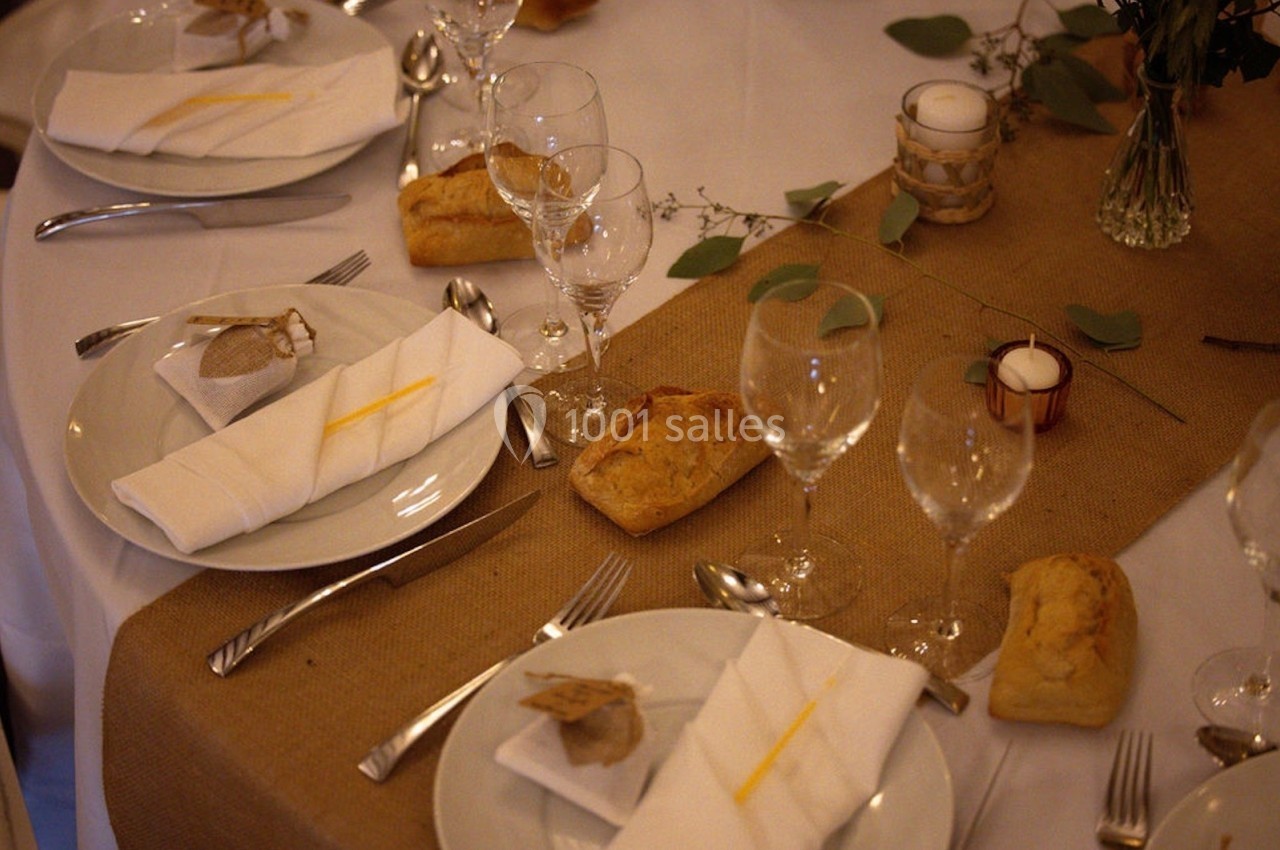 Table dressée avec nappes blanches, chemins de table en jute, verres, couverts, serviettes pliées et petits pains.