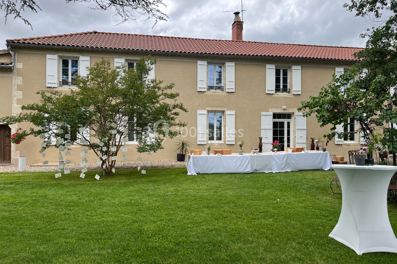 Grande maison en pierre avec volets blancs, pelouse verte au premier plan et table dressée pour un événement extérieur.