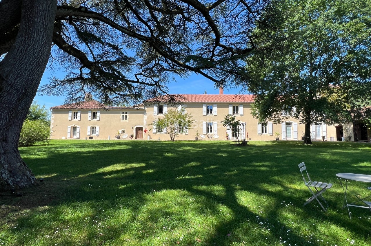 Grande maison en pierre avec toit rouge, entourée d'arbres et d'une pelouse ombragée, par une journée ensoleillée.