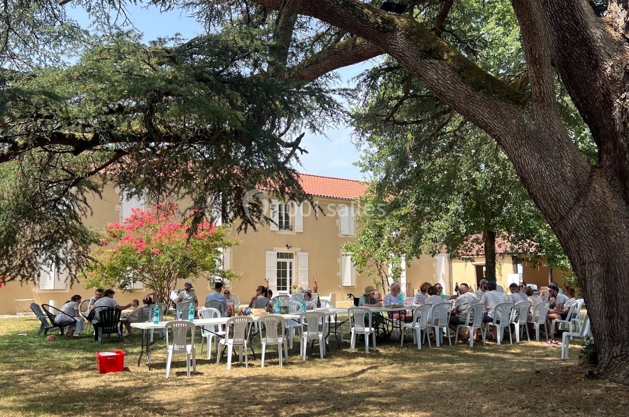 Groupe de personnes assises à des tables en plein air sous des arbres, devant une grande maison beige.