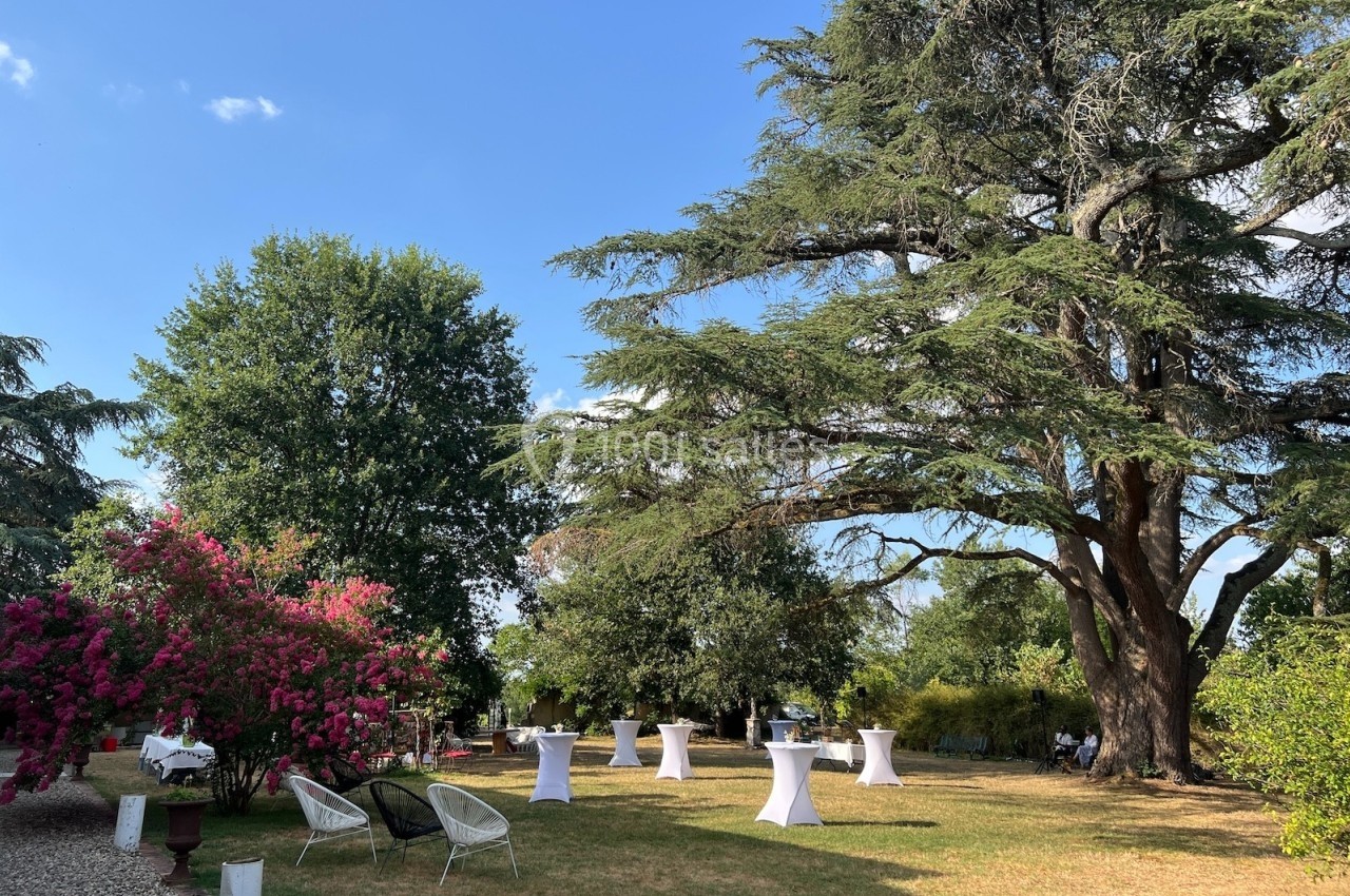 Jardin arboré avec tables hautes blanches, chaises et buissons fleuris sous un ciel bleu dégagé.