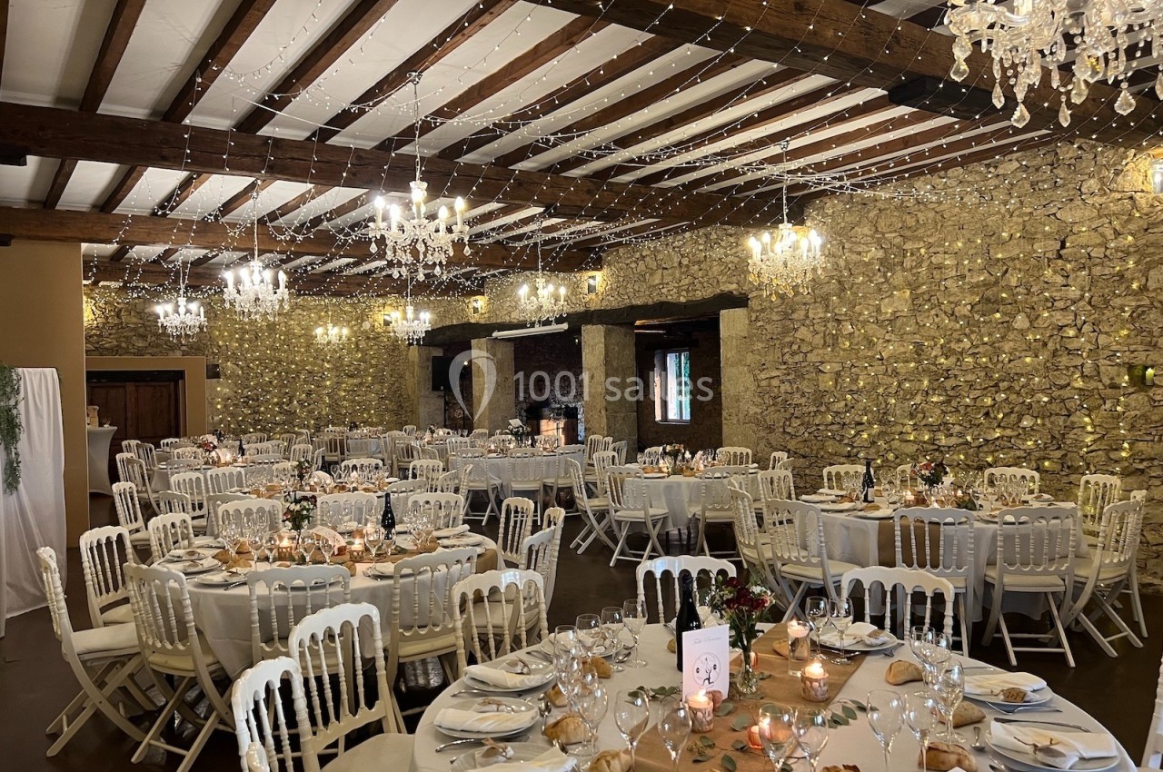Salle de réception décorée avec des tables rondes dressées, des chaises blanches et des guirlandes lumineuses suspendues.