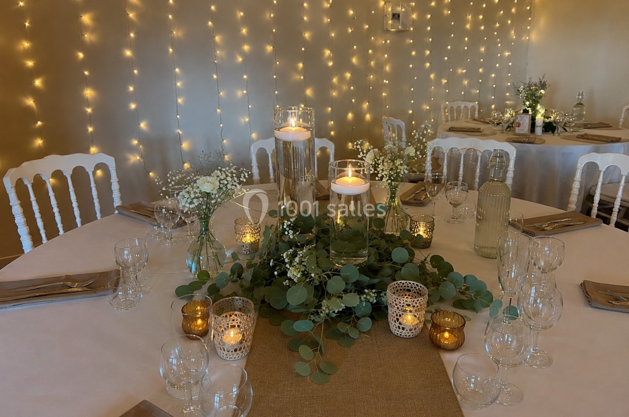 Table décorée pour un événement avec bougies, feuillage, nappes blanches et guirlandes lumineuses en arrière-plan.