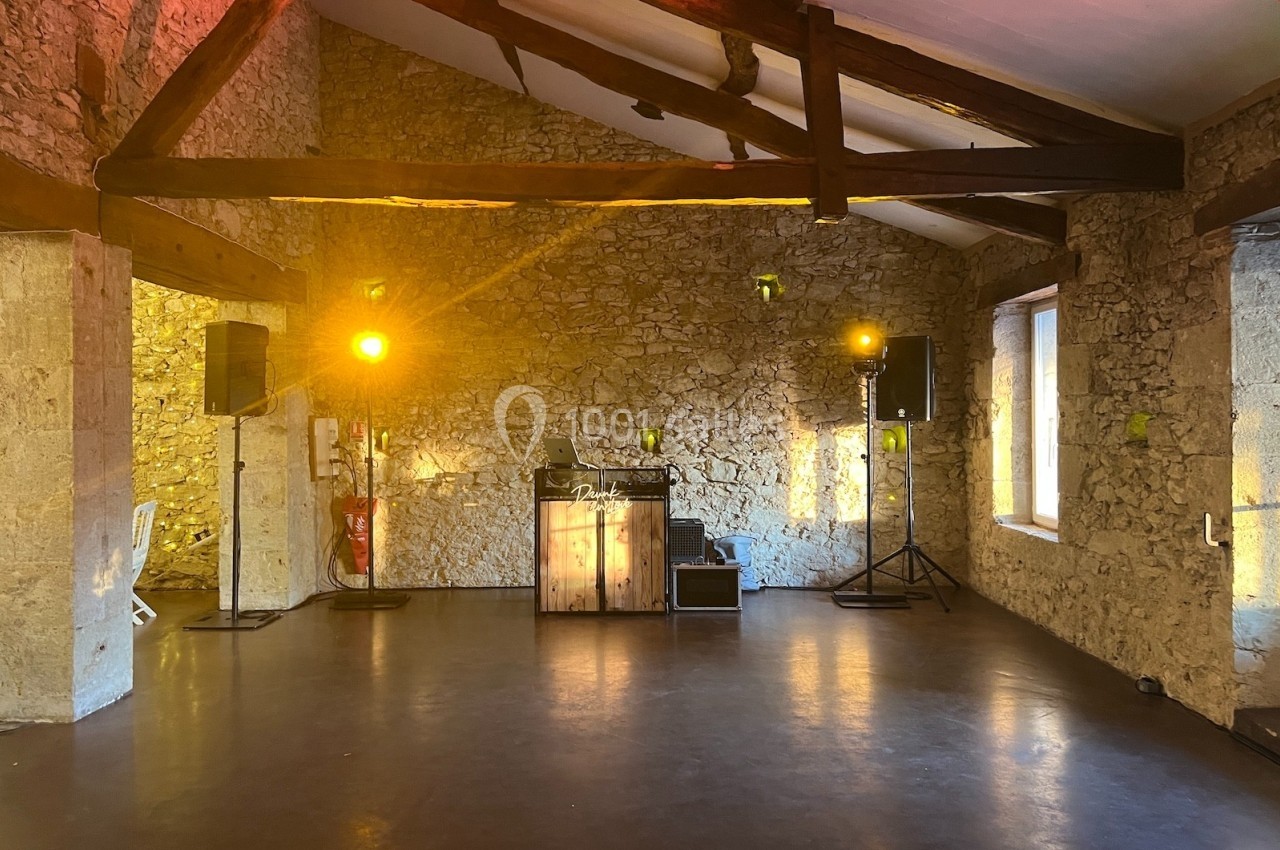 Salle en pierre avec poutres apparentes, éclairée par des lumières chaudes, équipée d'une table de DJ et de haut-parleurs.