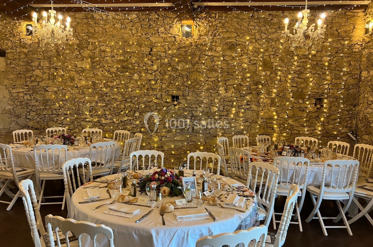 Salle de réception décorée avec des guirlandes lumineuses, des tables rondes dressées et des chaises blanches.