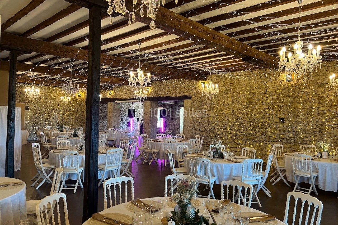 Salle de réception en pierre avec tables rondes dressées, chaises blanches et éclairage décoratif suspendu.