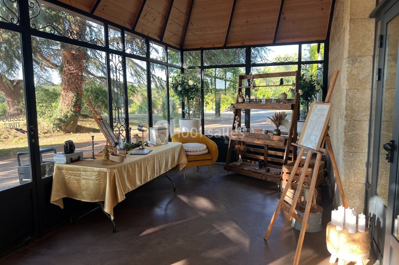 Salle lumineuse avec verrière, table décorée, étagères en bois, chevalet et vue sur un jardin arboré.