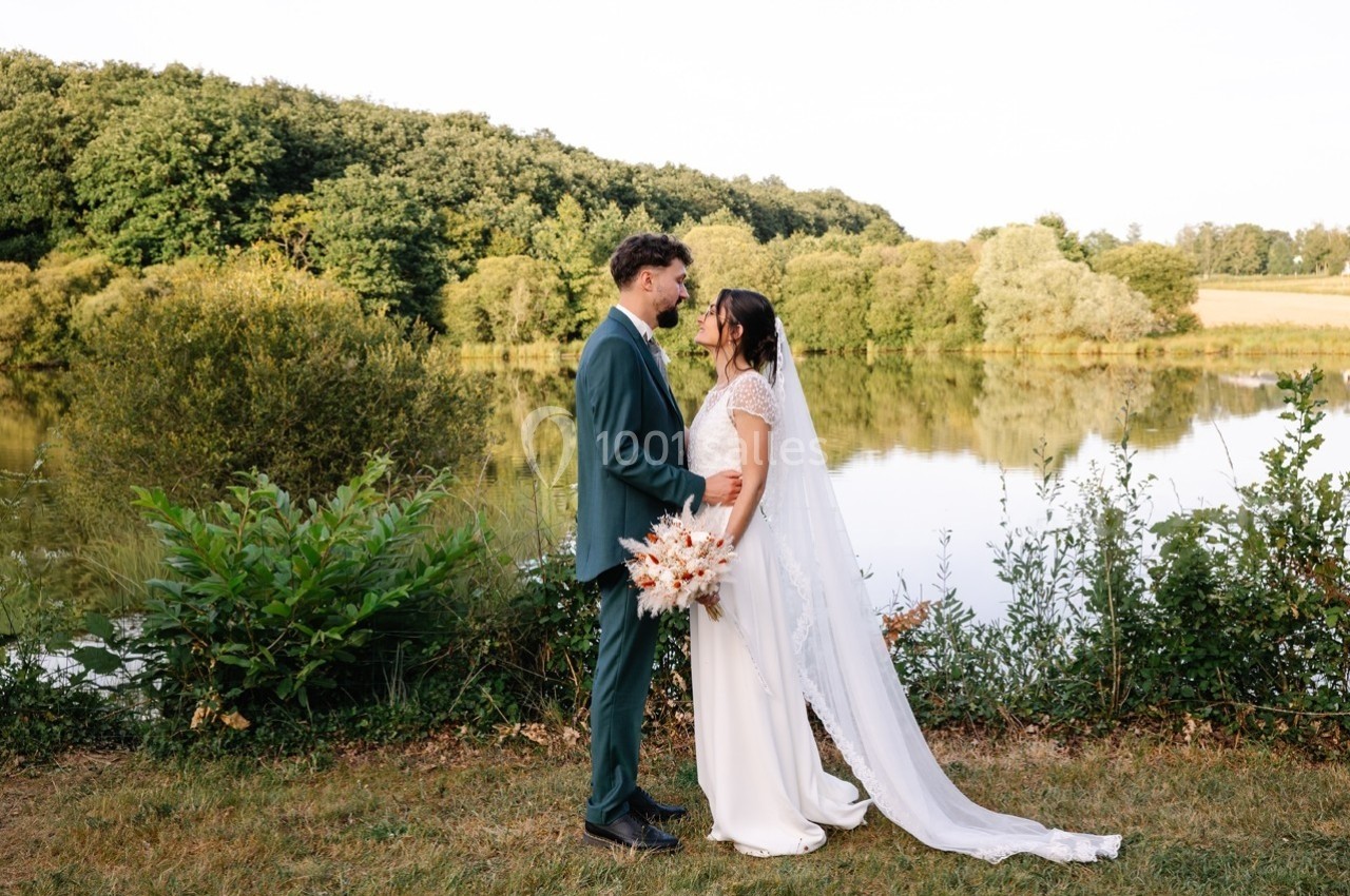 Un couple en tenue de mariage se tient face à face près d'un lac entouré de verdure.