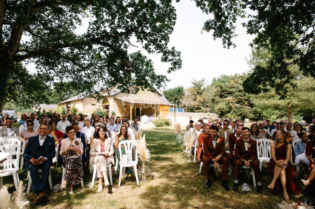 Invités assis en plein air lors d'une cérémonie, devant une maison en pierre entourée d'arbres.