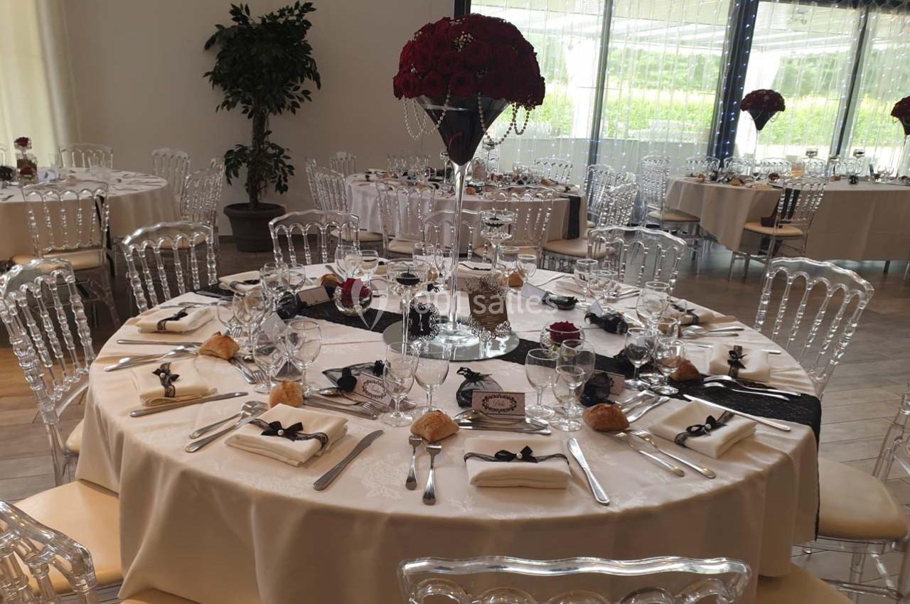 Table ronde élégamment dressée avec nappes blanches, serviettes pliées, verres, couverts et centre de table floral rouge.