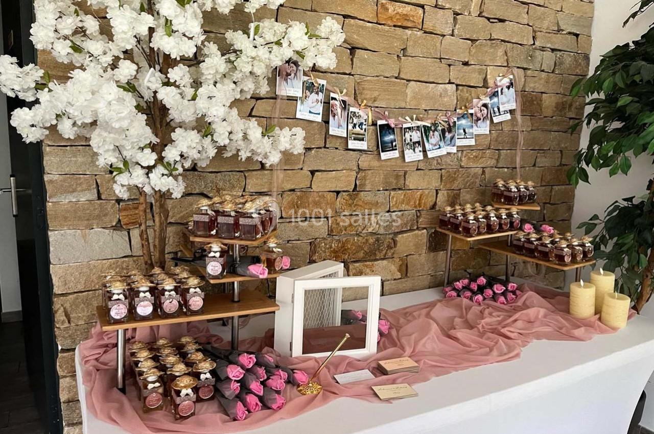 Table décorée avec des pots de confiture, fleurs roses, bougies et photos suspendues sur un mur en pierre.