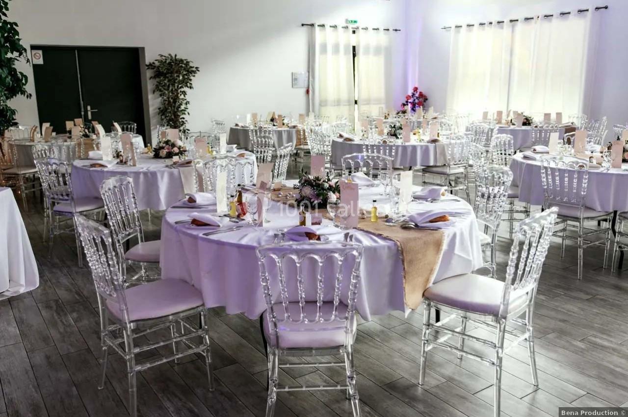 Salle de réception décorée avec des tables rondes, nappes blanches, chaises transparentes et centres de table fleuris.