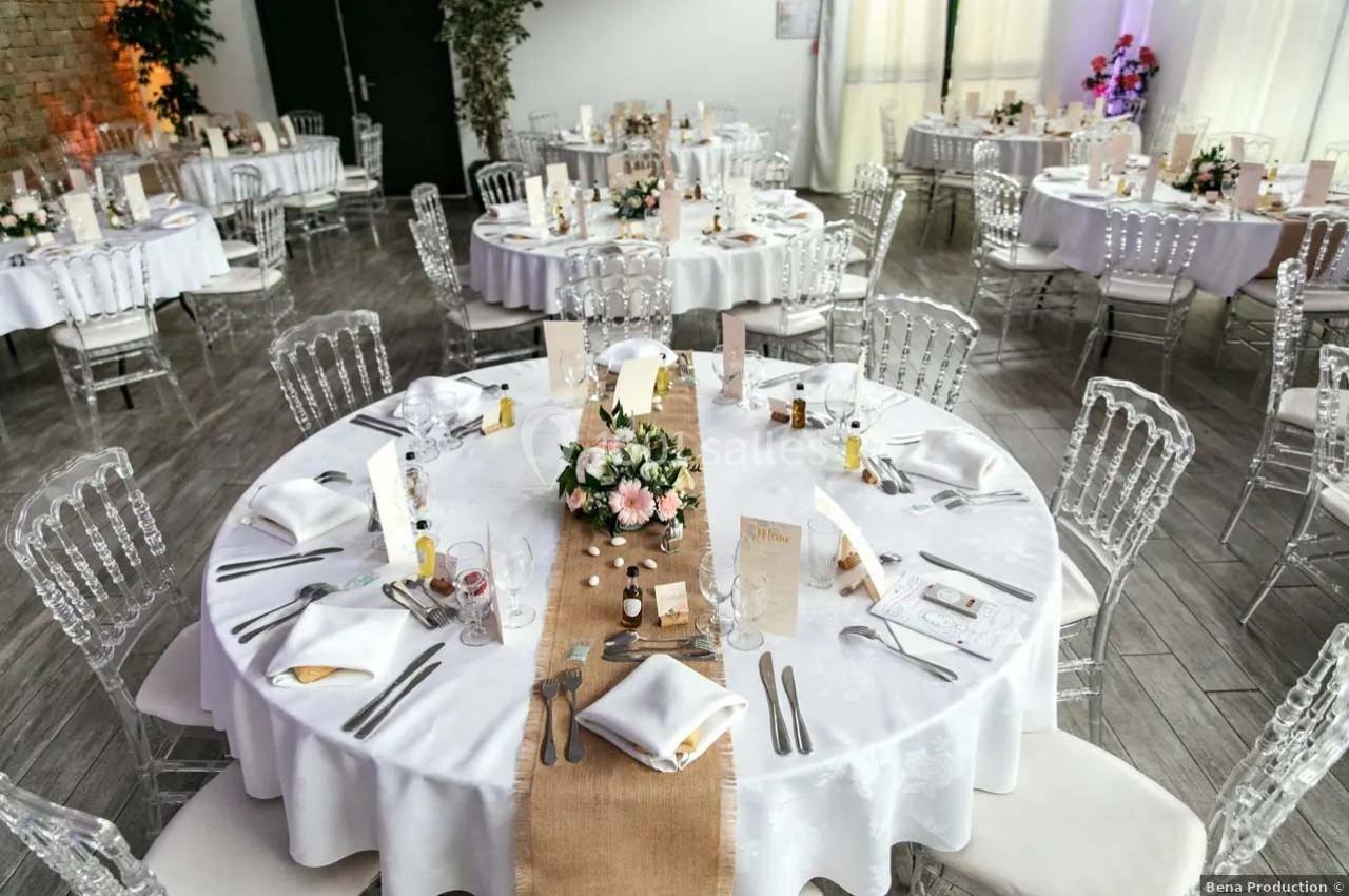 Salle de réception avec tables rondes dressées, nappes blanches, chaises transparentes et décorations florales.