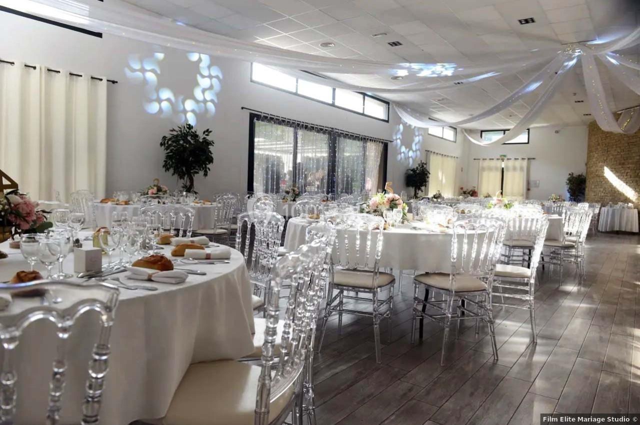 Salle de réception lumineuse décorée pour un événement, avec tables rondes dressées et chaises transparentes.