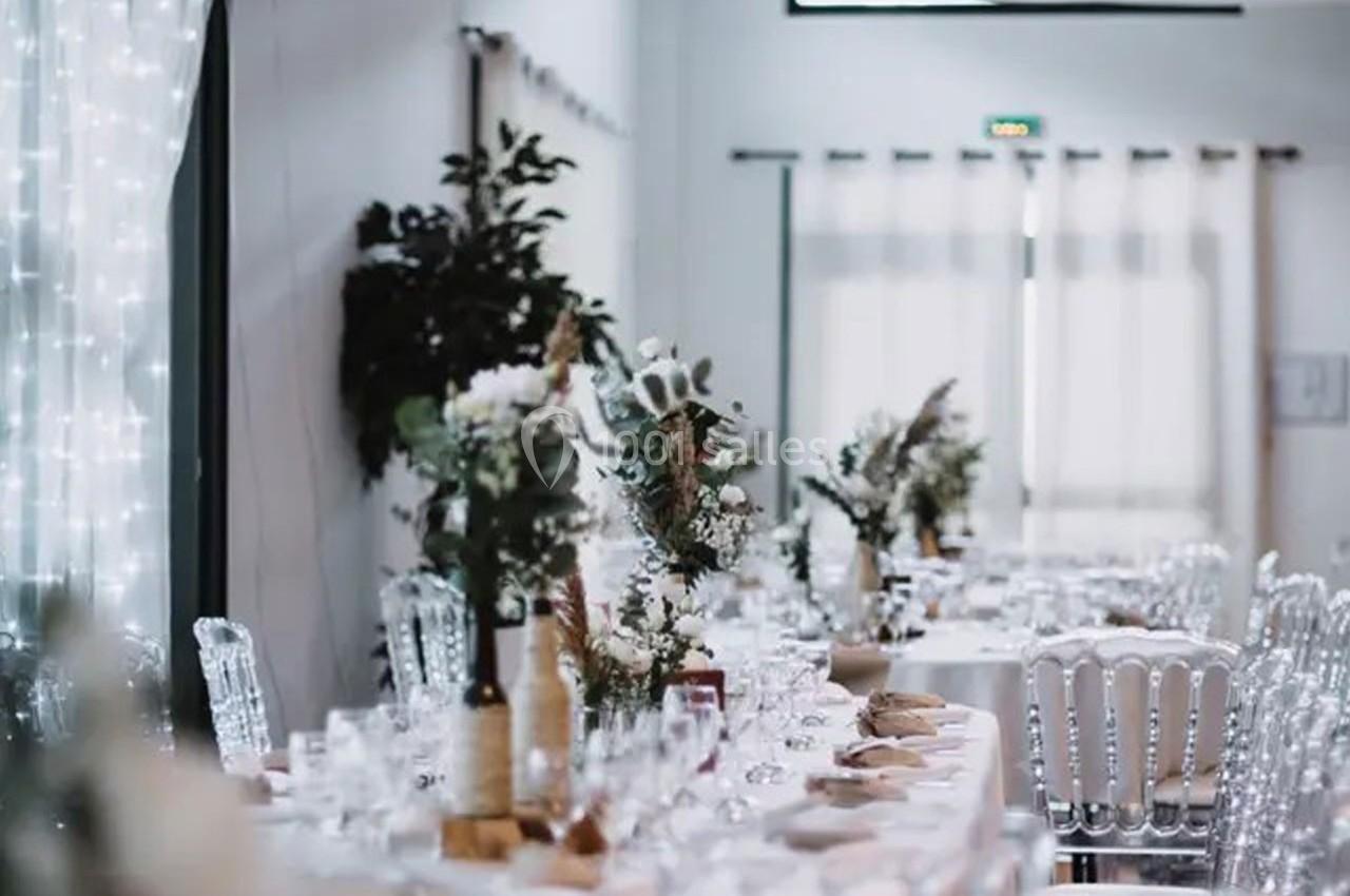 Salle décorée pour un événement avec tables dressées, chaises transparentes et arrangements floraux élégants.